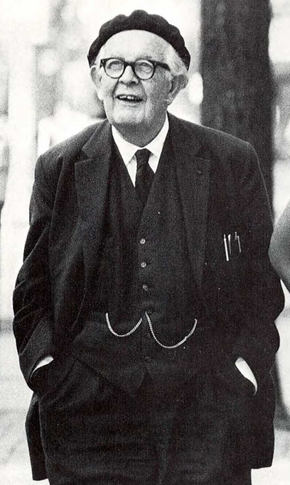 Jean Piaget sonríe de pie, vestido de traje y corbata. Tiene los anteojos puestos y una boina.