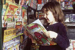 Una niña en un puesto de diarios y revistas. Sonríe entusiasmada mientras lee una revista.