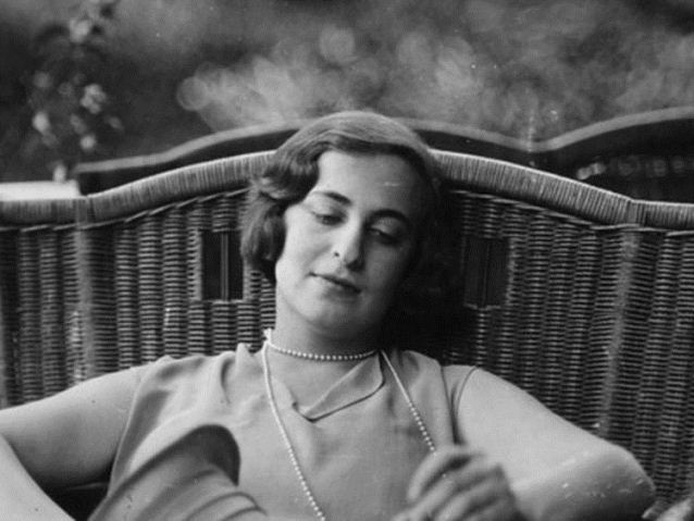Fotografía en blanco y negro de Silvina Ocampo joven, sentada en un sillón de mimbre al aire libre. Lleva el cabello corto y ondulado, un vestido claro sin mangas y un collar de perlas largo. Mira hacia abajo con expresión serena, mientras sostiene un objeto entre las manos.
