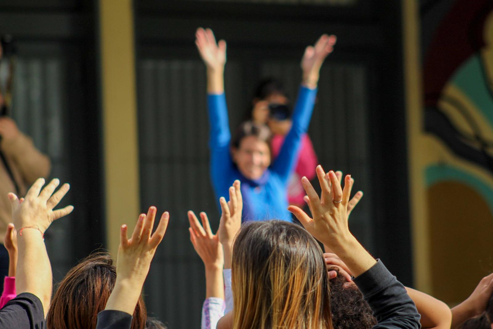 Grupo de personas con los brazos levantados. En primer plano se ven varias manos alzadas. Al fondo, desenfocada, hay una persona con camiseta azul que tiene los brazos en alto, y parece estar sonriendo. Detrás de ella hay otras personas.