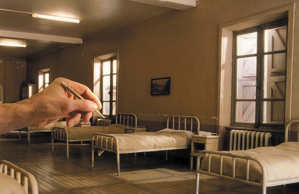 Una mano está ajustando un detalle en un dormitorio en miniatura. Hay varias camas de hierro y ventanas altas.