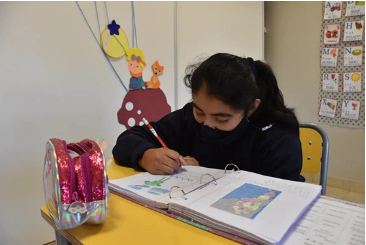 Niña con barbijo sentada en un pupitre, escribiendo con lápiz en su carpeta.