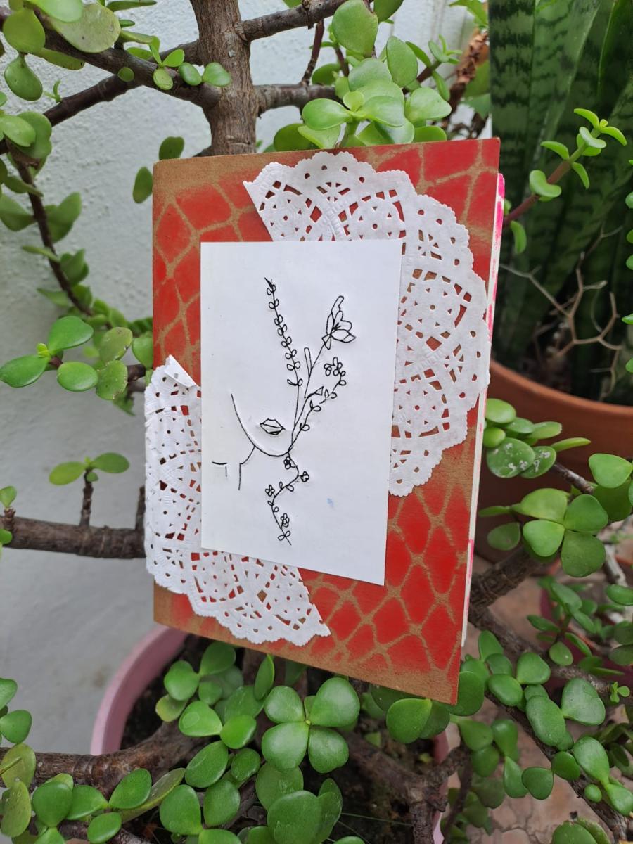 Un fanzine dispuesto entre las ramas de una planta. La tapa es roja y está decorada con blondas. En el centro, hay medio rostro dibujado, perfilado con hojas y flores.