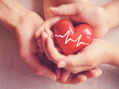Dos manos entrecruzadas sosteniendo un objeto rojo con forma de corazón. El objeto tiene dibujada en blanco la línea de un electrocardiograma.
