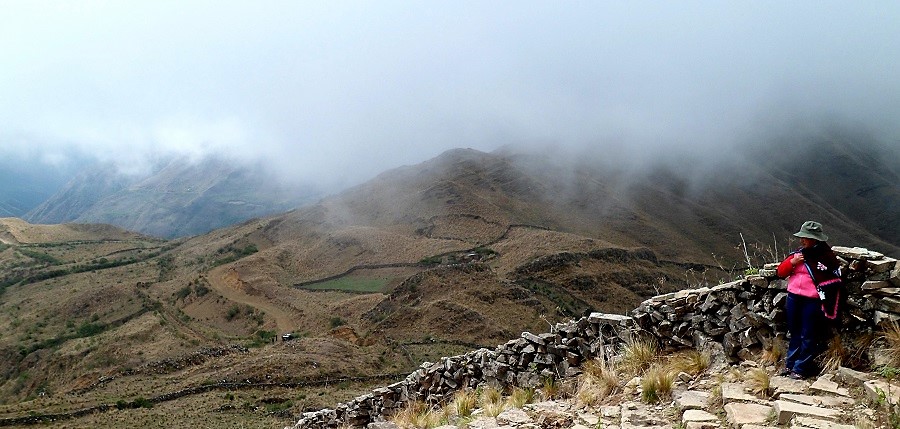 el sistema vial andino con montañas de fondo, una lugareña con sombrero, parada a la derecha de la imagen sobre el sendero