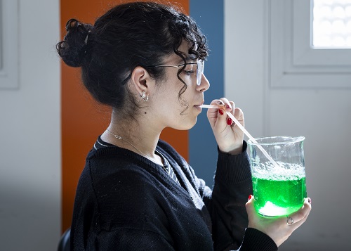 Una joven sopla con un sorbete en un frasco que tiene un líquido verde y hace burbujas.