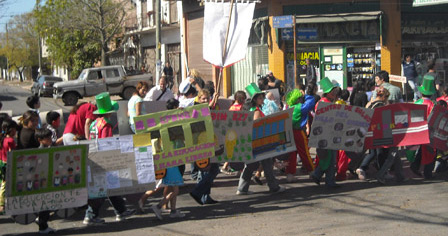 Chicos en fila caminan por una calle llevando pancartas y estandartes. Visten trajes de murga y algunos tienen galeras verdes.