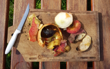una tabla de madera con un cuchillo apoyado y cáscaras de manzana, de batata, de palta y restos de cebolla.