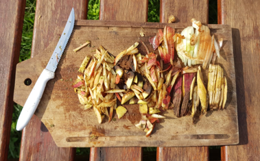 una tabla de madera con un cuchillo apoyado y pequeños pedacitos cortados de cáscaras de manzana, de batata, de palta y de restos de cebolla.
