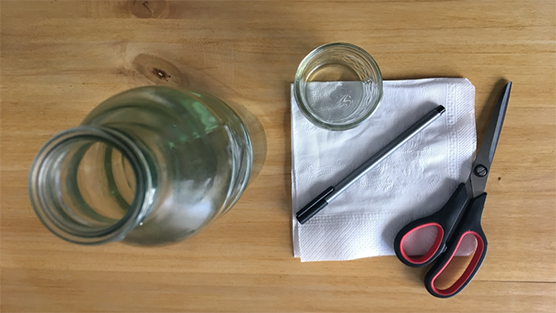 Una tijera, una servilleta de papel, una fibra negra, un frasco de vidrio vacío y una botella con agua.