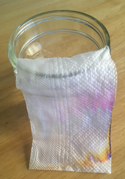 Un rectángulo de papel de servilleta con una mitad dentro y una por fuera de un frasco de vidrio, en el cual se observa un degradé de colores.