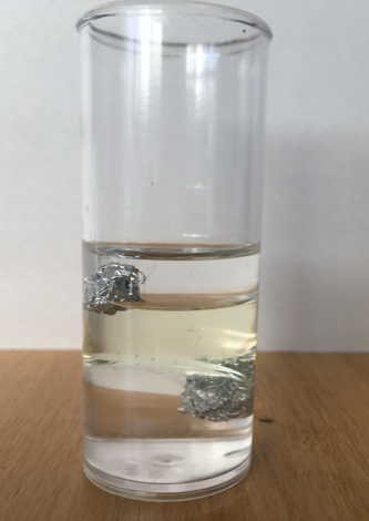 Vaso con agua, aceite y alcohol con las pelotitas de aluminio dentro.