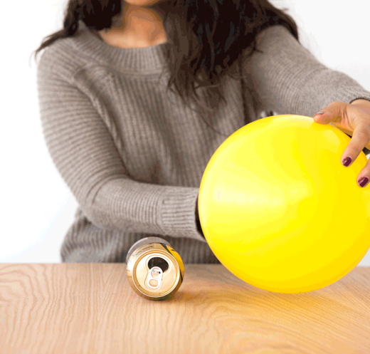 Una persona sostiene un globo inflado frente a una lata de aluminio colocada de forma horizontal sobre una mesa.