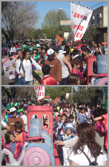 Festejo popular con personas adultas, niñas y niños. Algunos llevan sombrero y traje de murga. Hay estandartes de una escuela y un carro alegórico de una locomotora.