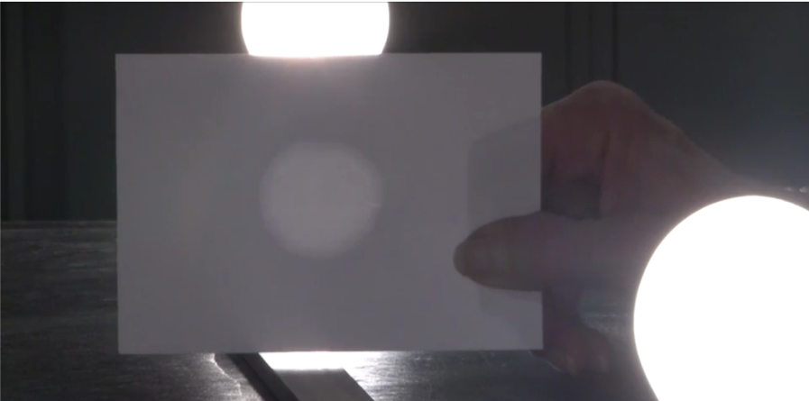 Experimento montado. Una mano sostiene el papel con la mancha de aceite en el centro a mitad de distancia entre dos foquitos encendidos. Fuente de la imágen: Oil Spot Photometer del Exploratorium Teacher Institute