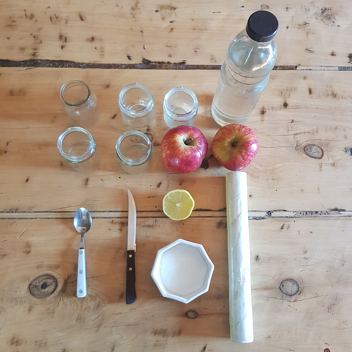  Una botella con agua, recipientes de vidrio, dos manzanas, medio limón, un cuchillo, una cucharita, un rollo de papel film y un recipiente con azúcar, sobre una mesa.