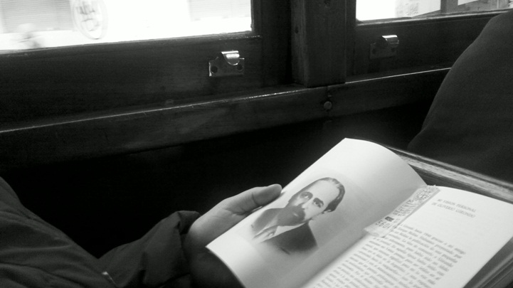 Una persona sostiene un libro de Oliverio Girondo en un tranvía, sentada junto a la ventanilla. En la página abierta hay un retrato del poeta. Fotografía en blanco y negro.