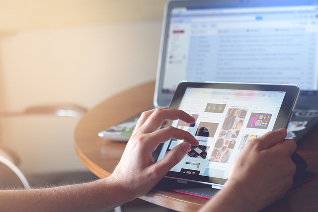 Las manos de una persona sosteniendo y operando una tableta con una computadora de fondo.