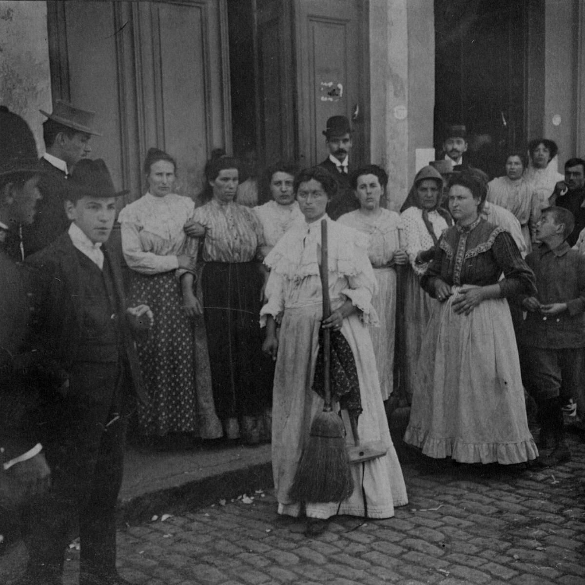 Mujeres y hombres con caras serias reunidos en una vereda. En el centro, una mujer con una escoba en la mano.