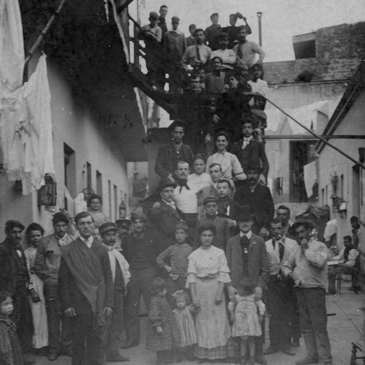 Patio de un conventillo. De pie, hombres, mujeres y niñes miran a cámara desde la planta baja y la escalera que va al primer piso.