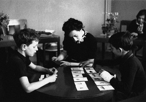 María Montessori y dos niños mirando unas fichas sobre una mesa.