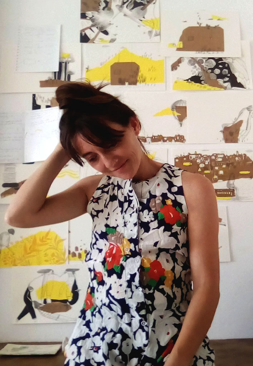 Nadia Romero Marchesini sonríe mirando hacia abajo. Detrás, hay varias ilustraciones suyas en amarillo, negro y marrón colgadas en una pared.