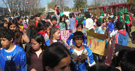 Multitud de niñas y niños en pleno festejo. Algunos tienen trajes de murga de color azul y otros, más atrás, traje y galera verdes. Al fondo, hay árboles.
