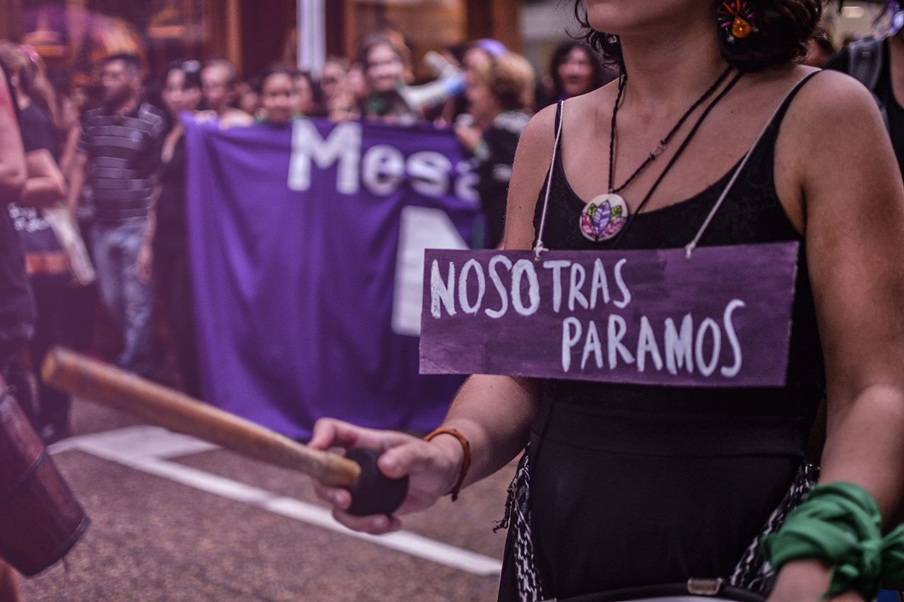 Fotografía color tomada durante una marcha por el 8M. Se ven personas fuera de foco al fondo y en primer plano una mujer que lleva colgado al cuello un cartel con el mensaje: nosotras paramos.