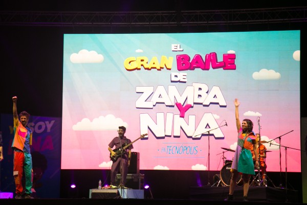 Tres jóvenes en un escenario. Dos alzan un brazo. De fondo, hay una pantalla gigante que dice: «El gran baile de Zamba y Nina».