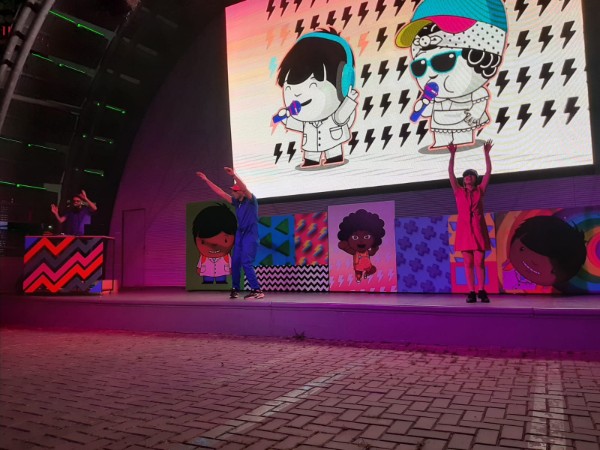 En un escenario, bailan un hombre y una mujer vestidos con ropas de color. Detrás, hay una pantalla enorme con los personajes de Zamba y Nina.