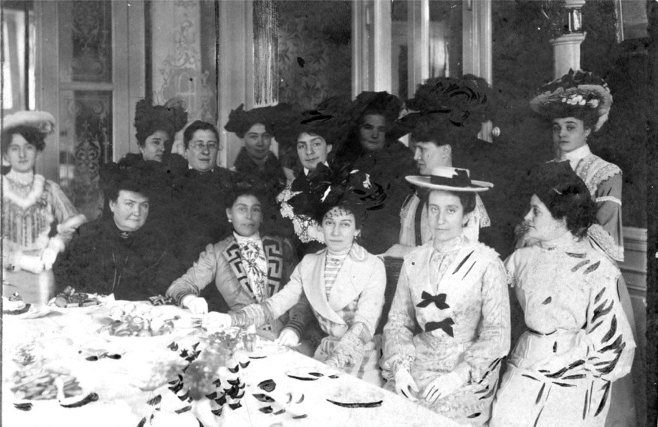 Lola Mora y un grupo de mujeres junto a una mesa servida. Algunas están paradas; otras, sentadas. Llevan vestidos y capelinas.