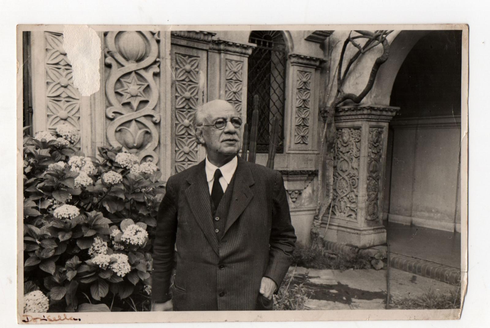 Ricardo Rojas en su jardín, de pie junto a hortensias y, de fondo, una pared con relieves ornamentales de su casa.