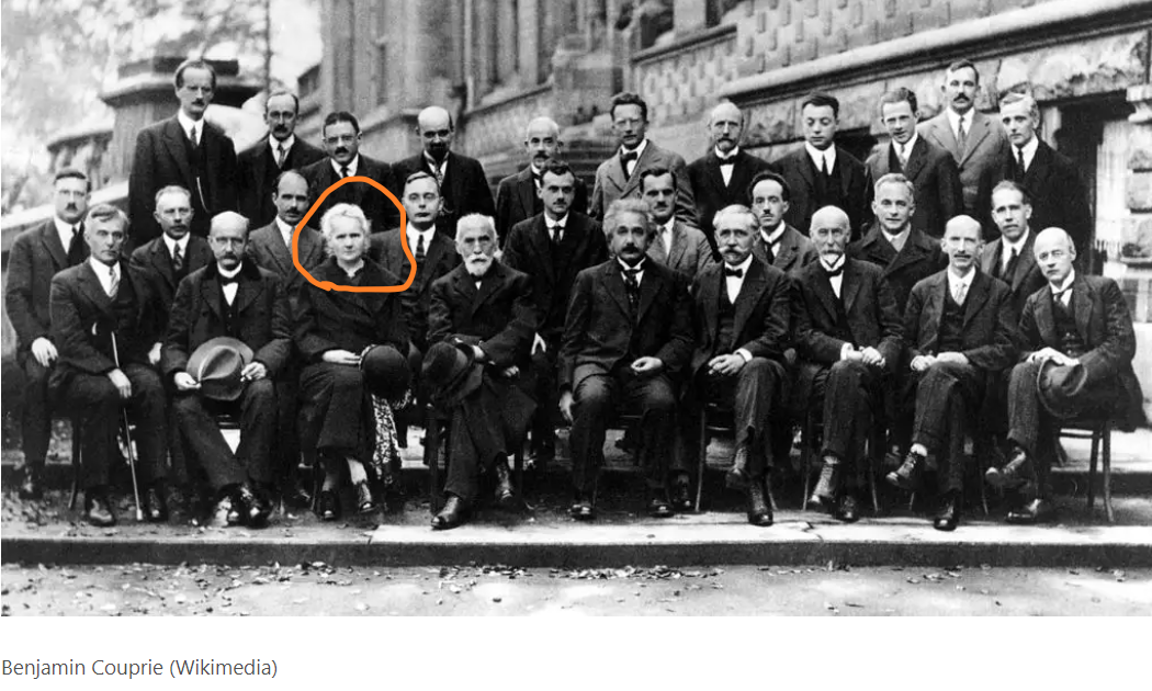 Grupo de hombres trajeados, algunos de pie y otros sentados, entre los que se encuentra en primera fila Marie Curie. Es la única mujer del grupo.