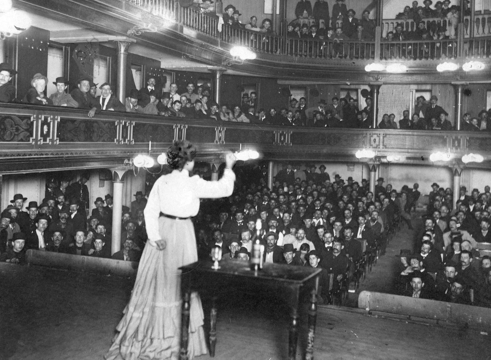 En un escenario, una mujer de pie alza el puño; viste blusa y falda. Solo hay hombres en el público.
